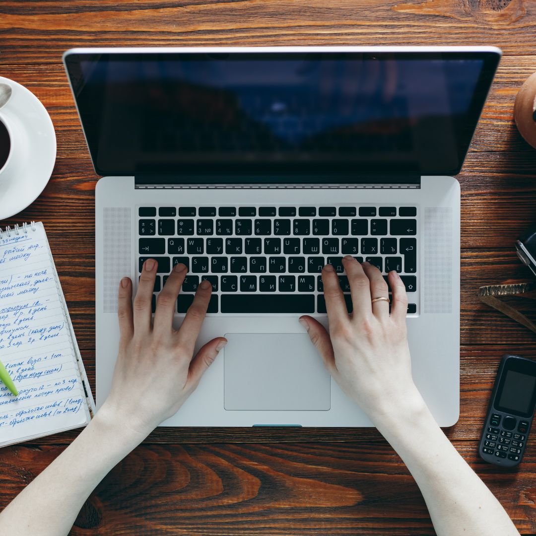 A person typing on a laptop, accompanied by a cup of coffee and an open notebook on a desk.