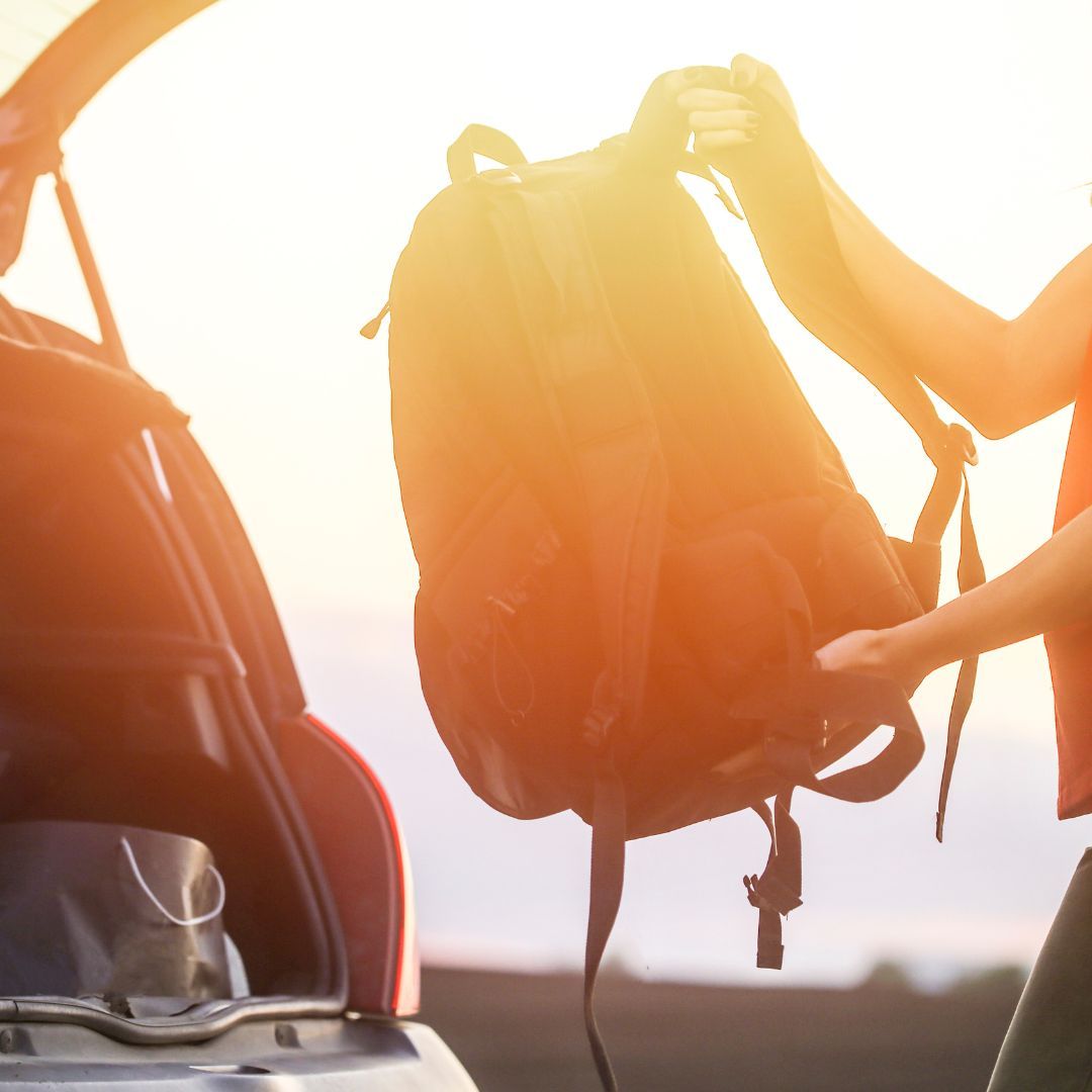 A woman stands at the back of a car, holding a backpack with a focused expression on her face.
