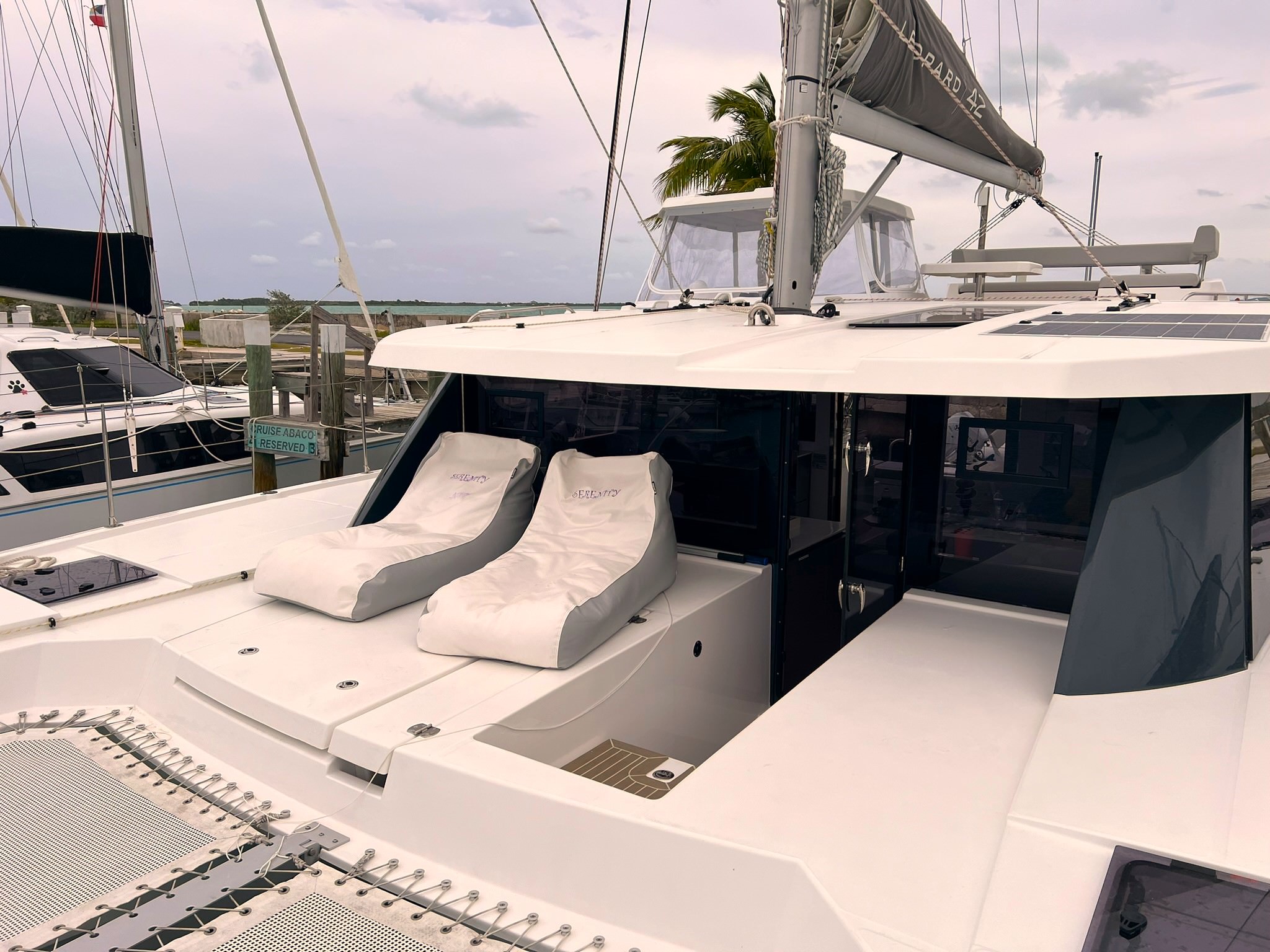 Serenity – Exterior 01 A catamaran featuring a pristine white deck adorned with comfortable chairs, set against a serene water backdrop.