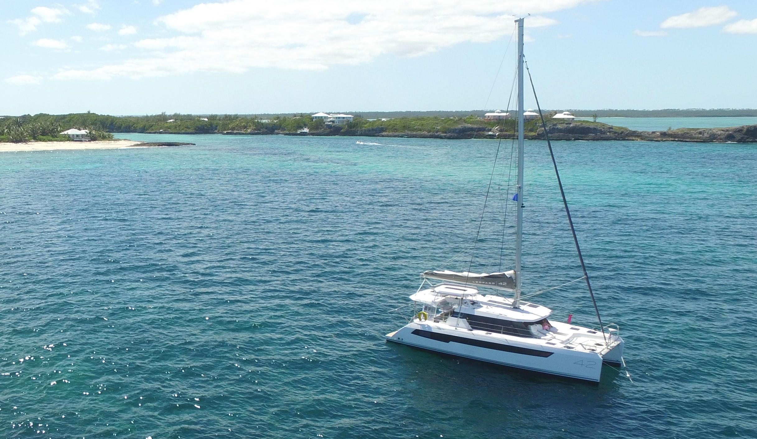 Serenity – Exterior 05 A catamaran gliding through the ocean waters, with a picturesque beach in the background under a clear blue sky.