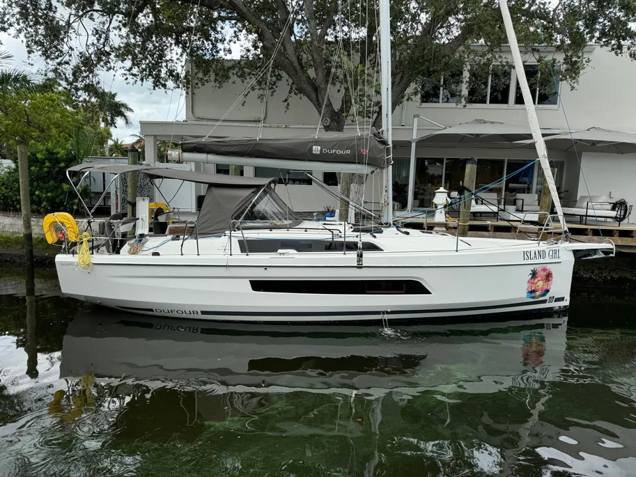 A sailboat is docked at a pier in front of a house, surrounded by calm water and a clear blue sky.