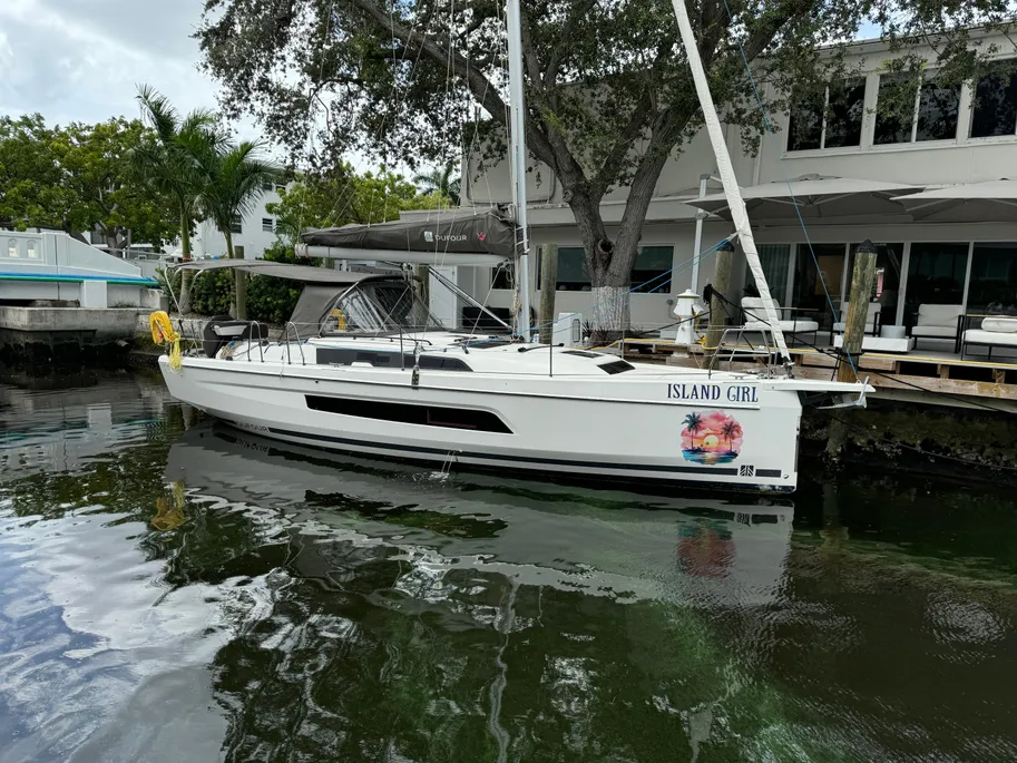 A sailboat is moored at a dock in front of a house, with serene water reflecting the clear sky above.