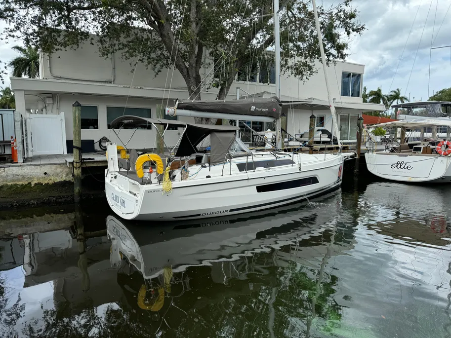 A sailboat is docked beside a house at a marina, with calm water reflecting the surroundings.